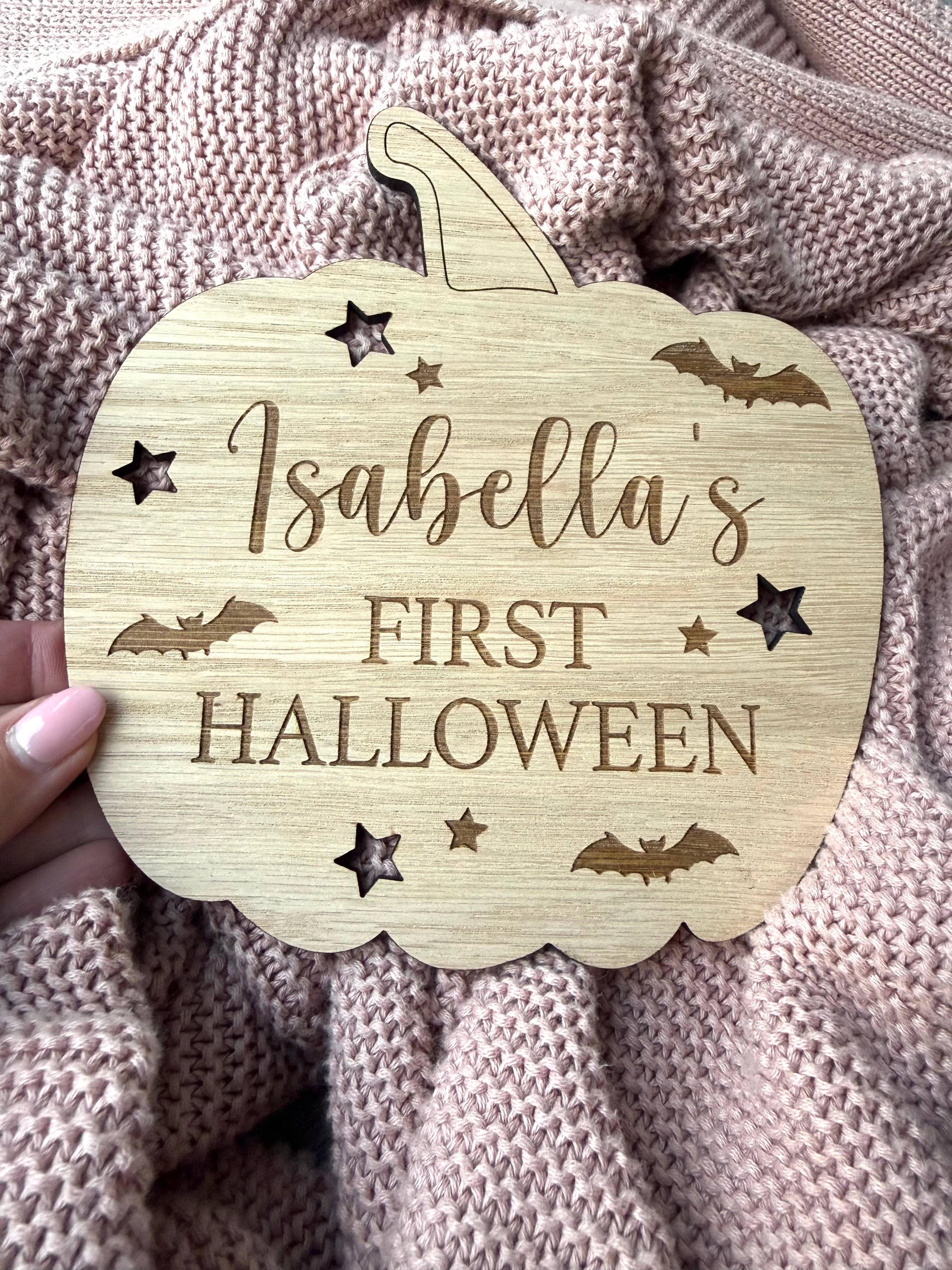 Wooden pumpkin-shaped plaque with 'Isabella's First Halloween' text on a textured fabric background